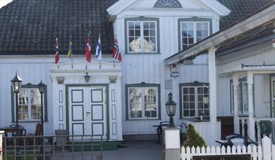 gammel hvit trebygning med blå vinduskarmer, smijernsport og flagg