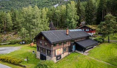 flyfoto av Hillestad galleri, en tømmerbygning omgitt av grønt gress og skog.