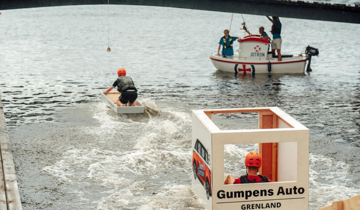 tre hjemmelagde båter på sjøen, hvite med rødt og oransje farger, to mennesker med oransje hjelmer, tre menn i båt med mørke skjorter