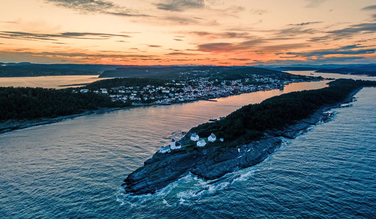 øy med hvitt fyrtårn og fire hvite hus rundt, fjell og skog med by i bakgrunn med hvite hus, oransje himmel og grå skyer