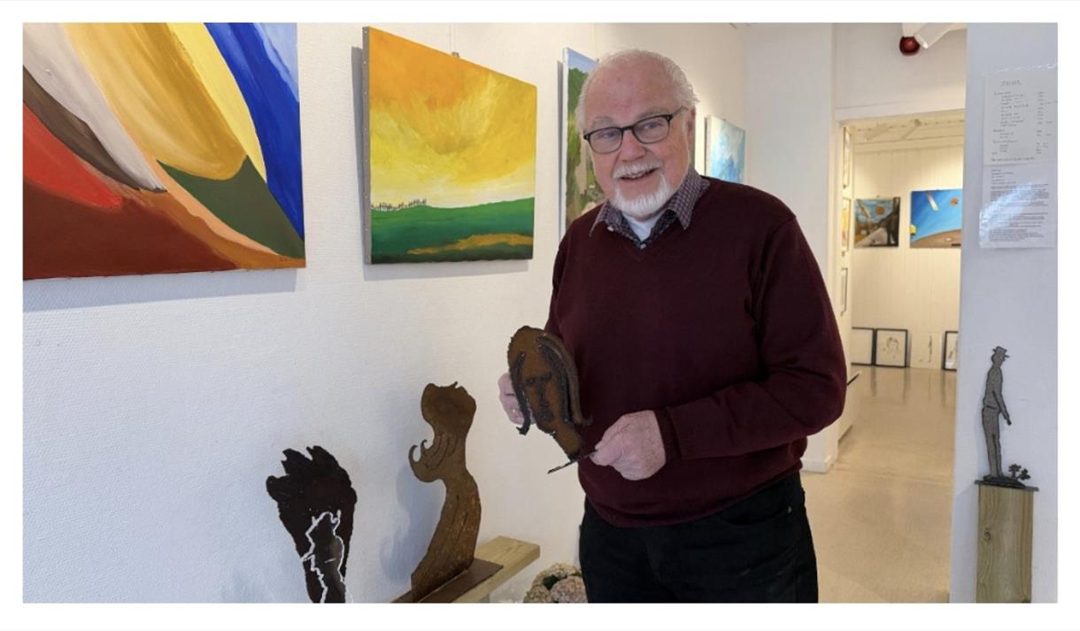 mann med hvitt hår, briller og skjegg, foran bilder med mye farger