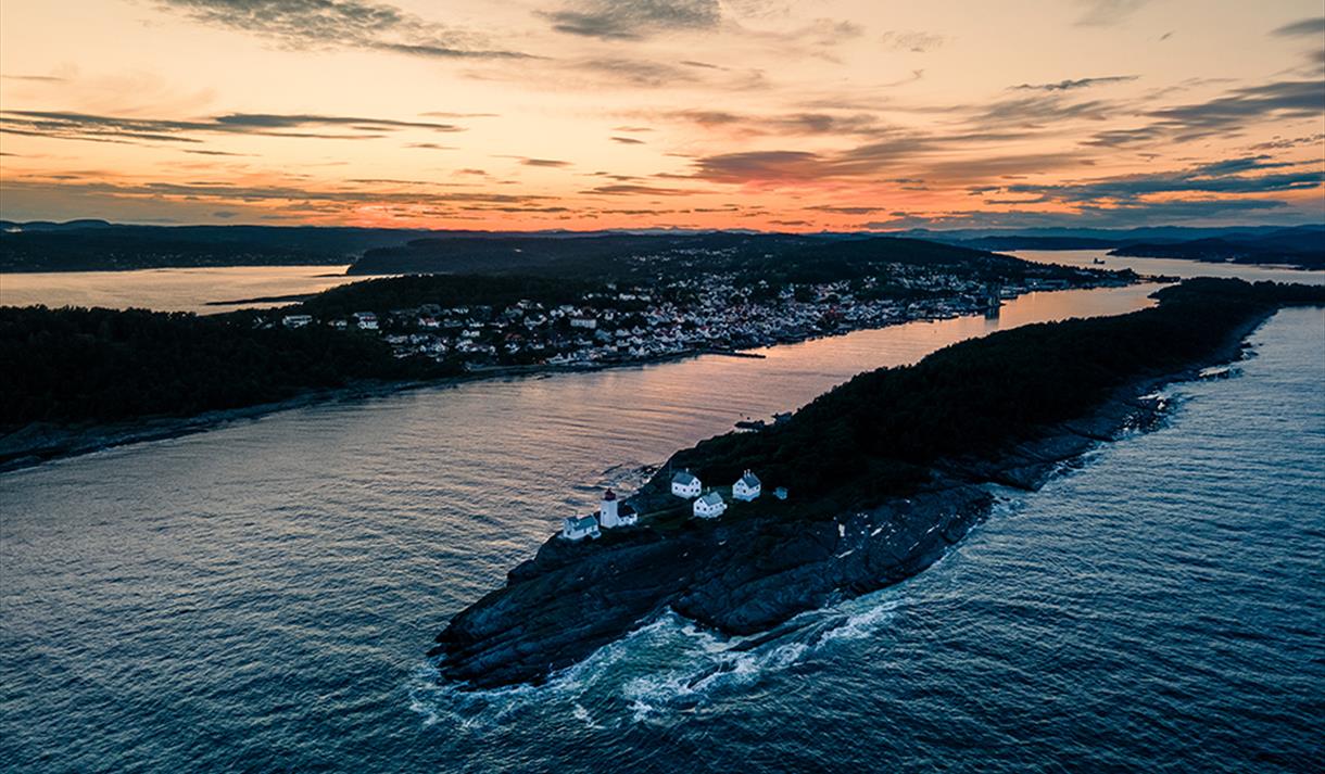 øy med hvitt fyrtårn og fire hvite hus rundt, fjell og skog med by i bakgrunn med hvite hus, oransje himmel og grå skyer