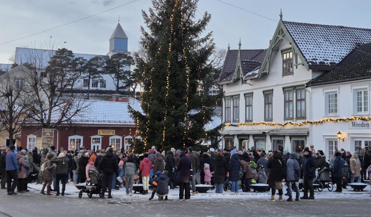 ulegran med lys og mennesker som ser på , kirke i bakgrunnen og rødt og hvitt trehus med julelys