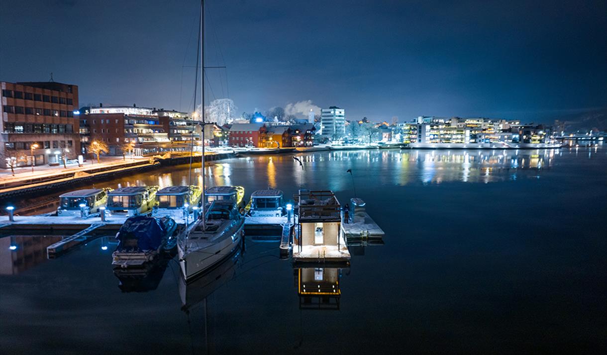 sauna liggene ved elva, med båter på brygge. blokker med lys i bakgrunnen