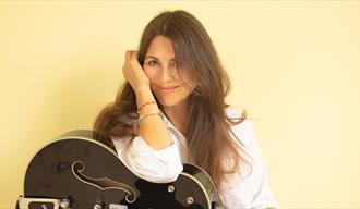 dame med langt brunrt hår i hvit bluse, sort gitar og gul bakgrnn