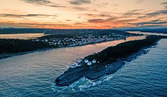 øy med hvitt fyrtårn og fire hvite hus rundt, fjell og skog med by i bakgrunn med hvite hus, oransje himmel og grå skyer