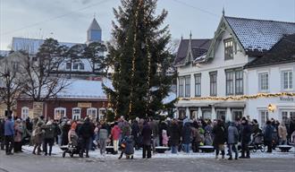 ulegran med lys og mennesker som ser på , kirke i bakgrunnen og rødt og hvitt trehus med julelys