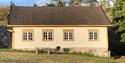 gammelt beige trehus med oransje karmer rundt smårutete vinduer. Gressplen med trær i bakgrunnen og sittebenk