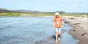 gutt med hvit hatt og orange tskjorte i shorts gående på sandstrand