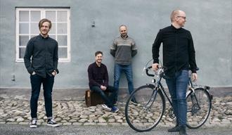 Hva skjer i Porsgrunn - Barnespeilet Mandarinsaft. Bildet: 4 menn som står foran den vegg, den ene med sykkel