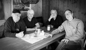fire menn sittene rundt et bord på en hytte med trepanel. kaffekopper og duk på bordet