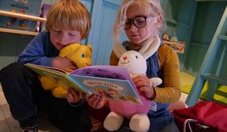 Karsten og Petra lager museum. 2 små barn, en gutt og en jente, som sitter med hver sin bambse og leser i en bok